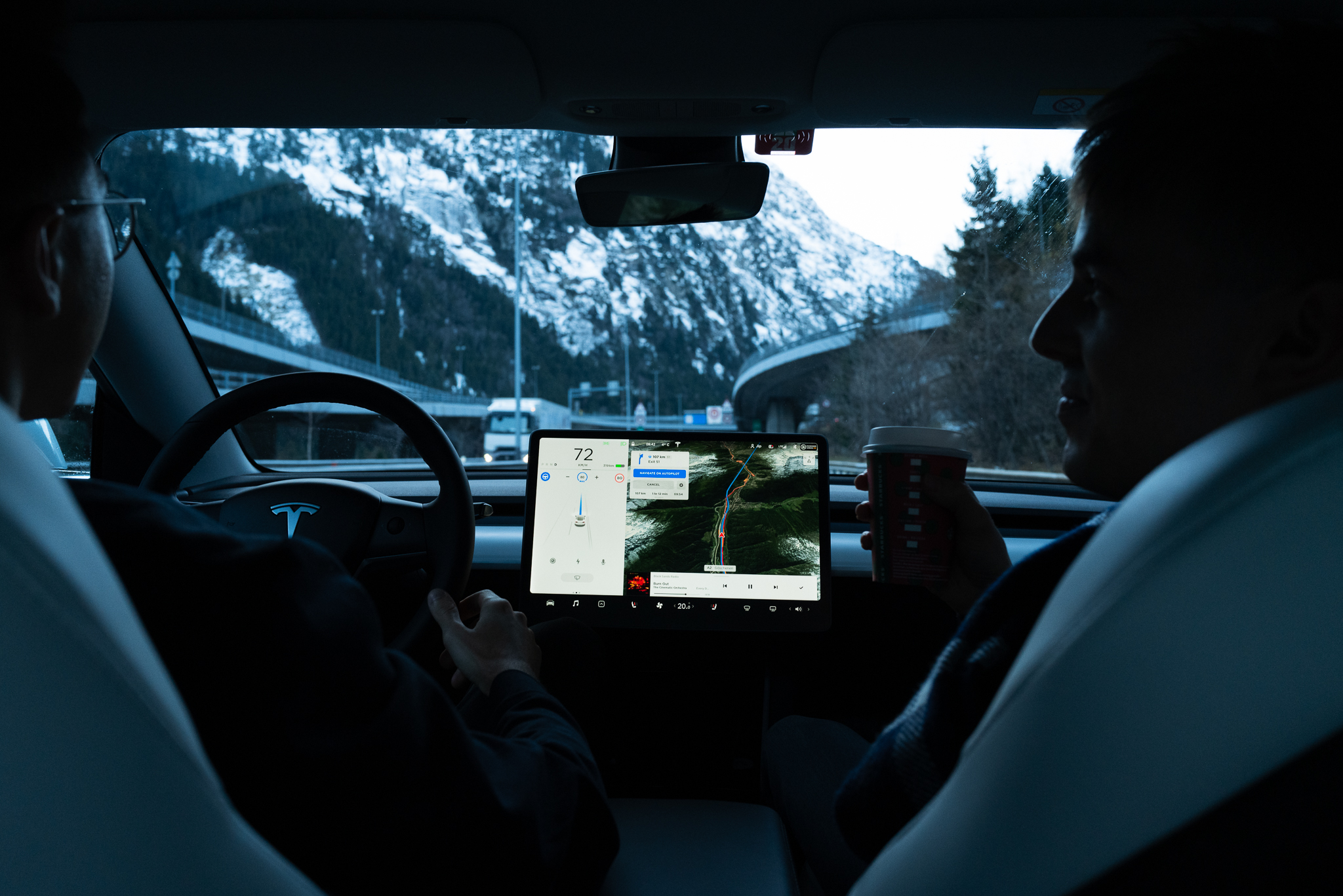 View within the Tesla from the back seat. Passengers drink a coffee. Snowy mountain landscape in Switzerland.