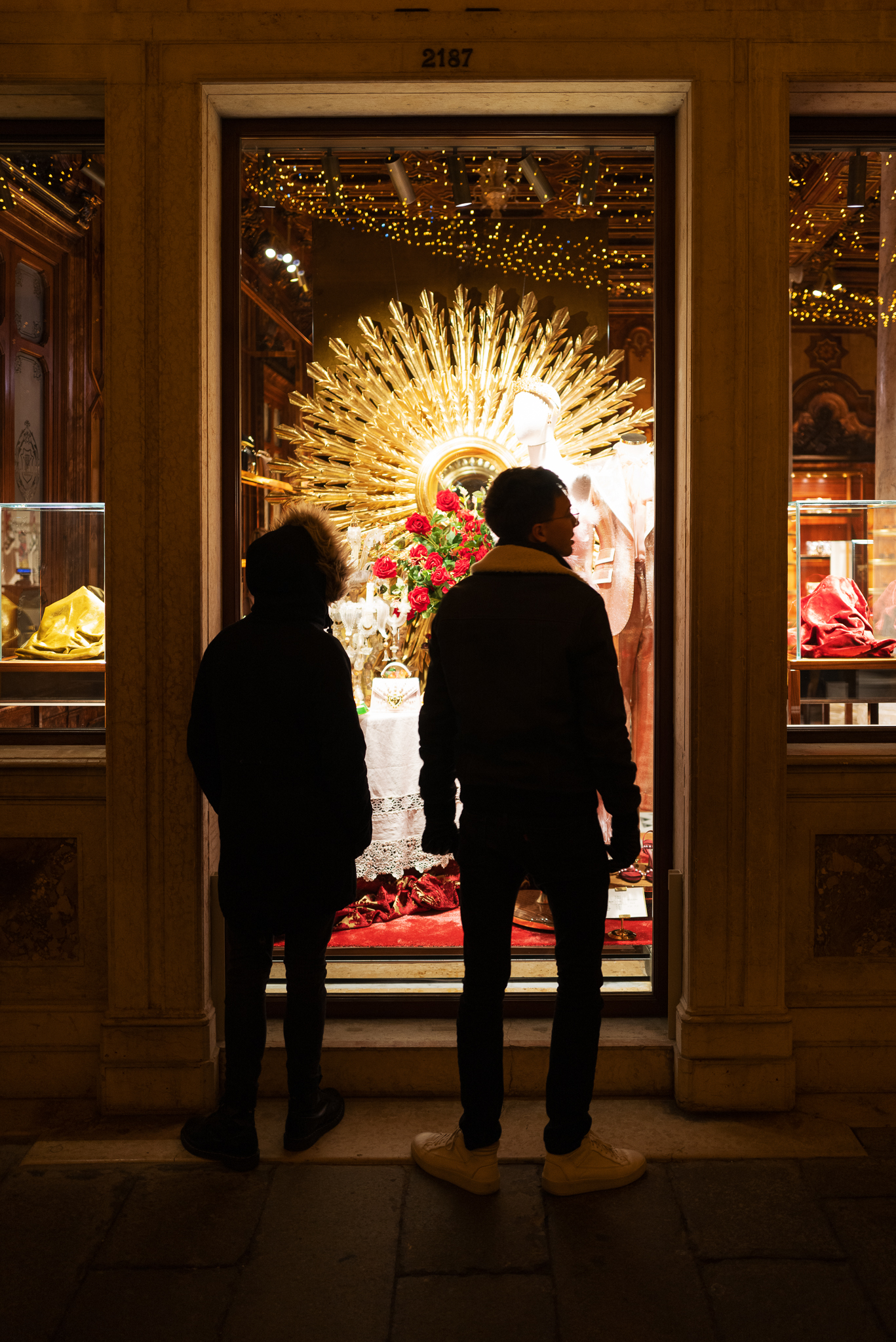 Two friends standing in front of the window of a beautifully decorated clothing shop.