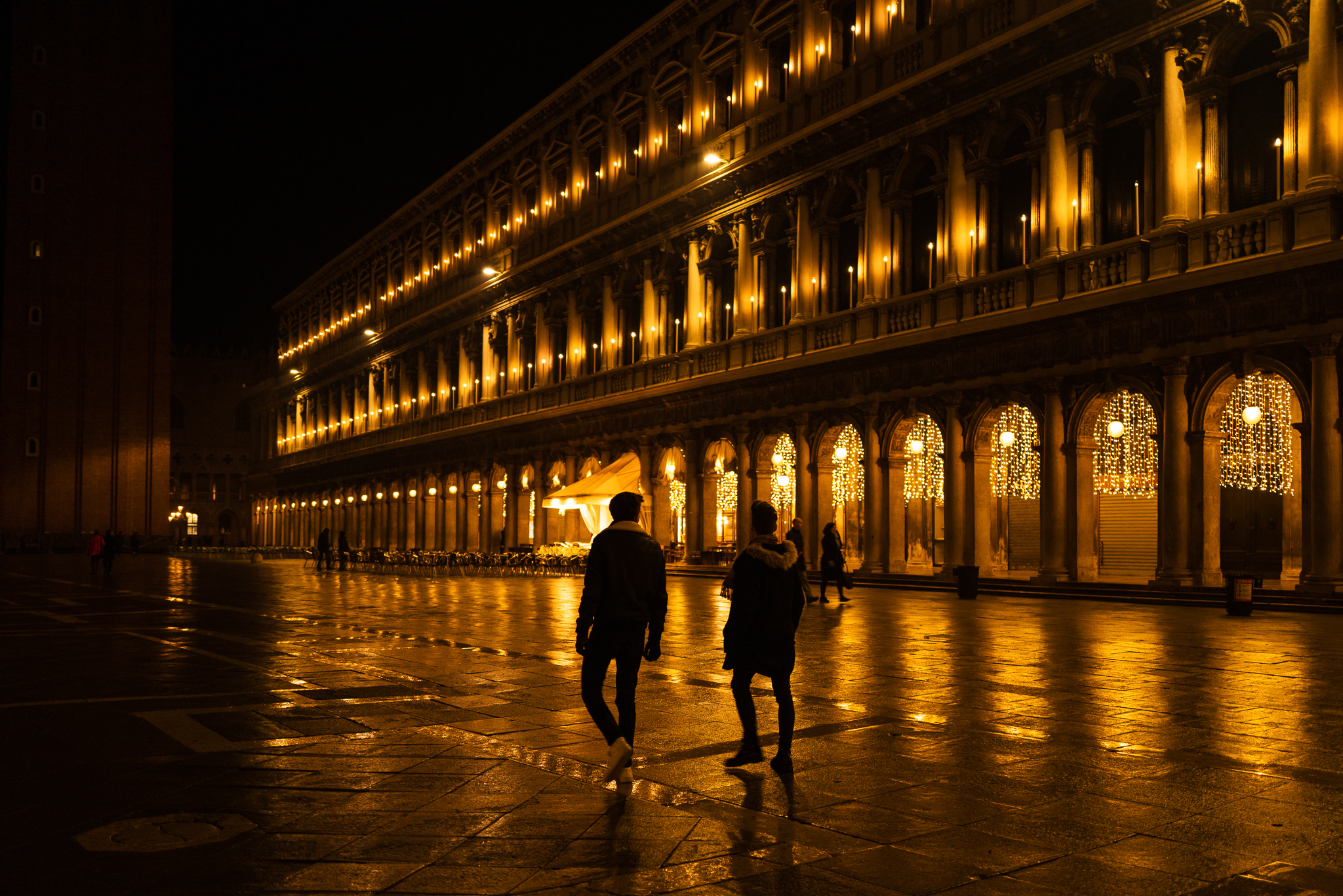 Two friends have a walk on San Marco square that is lit with golden Christmas decoration. There are few other people.