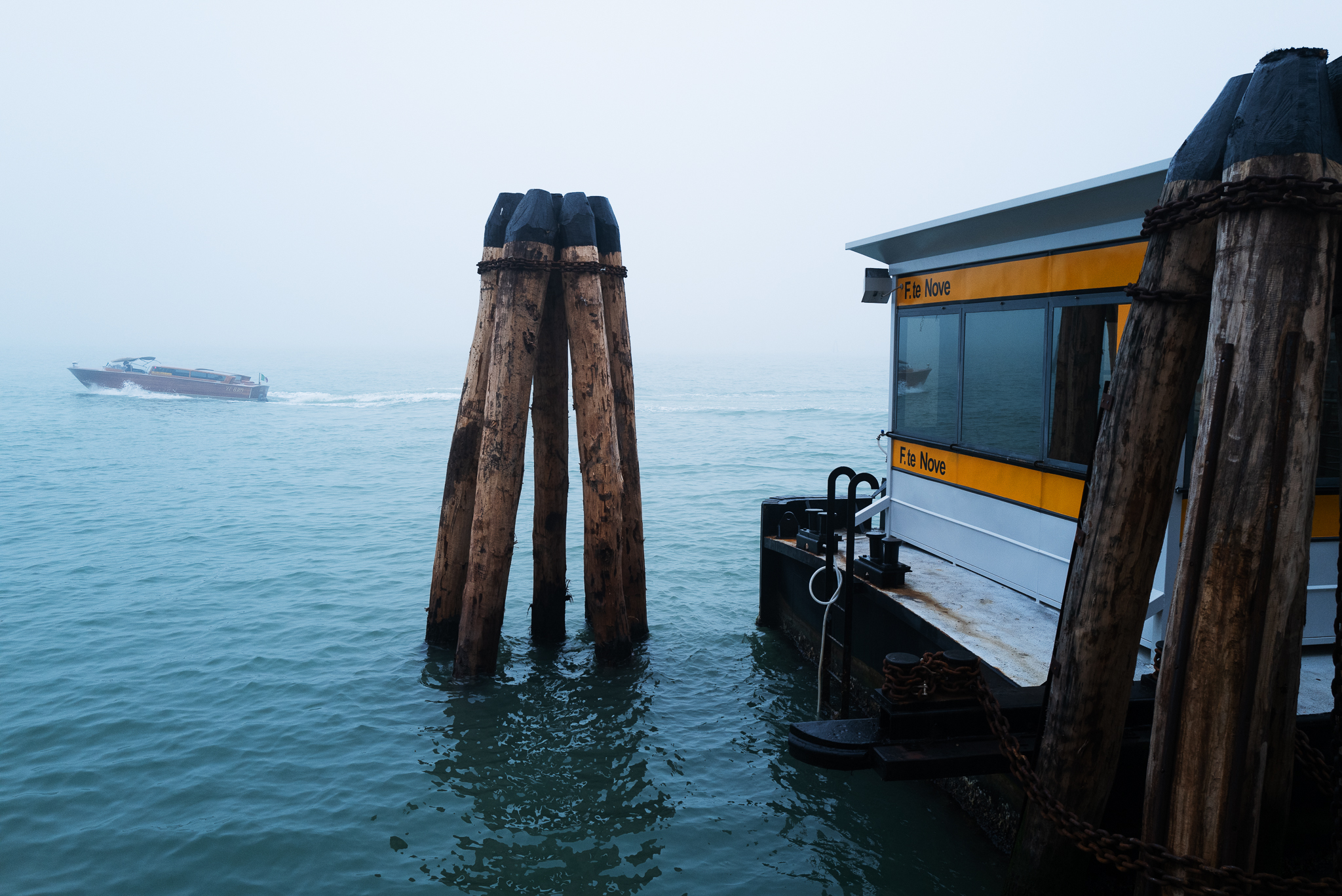 A Venice water taxi station and a boat passing by. It's foggy.